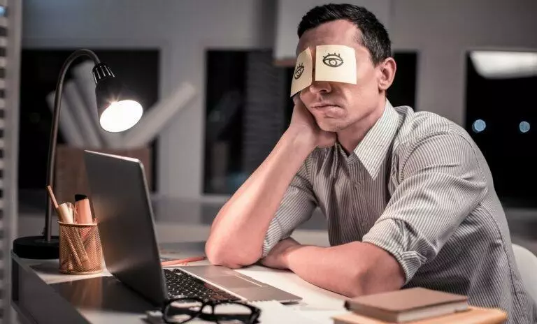 tired man on desk with paper stuck in eyes