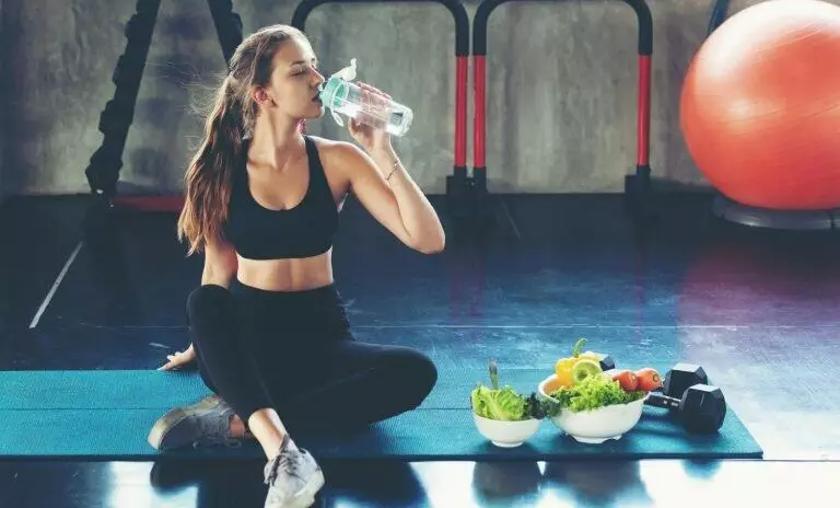 mage of a woman drinking water with bowls of vegetables beside her after a workout