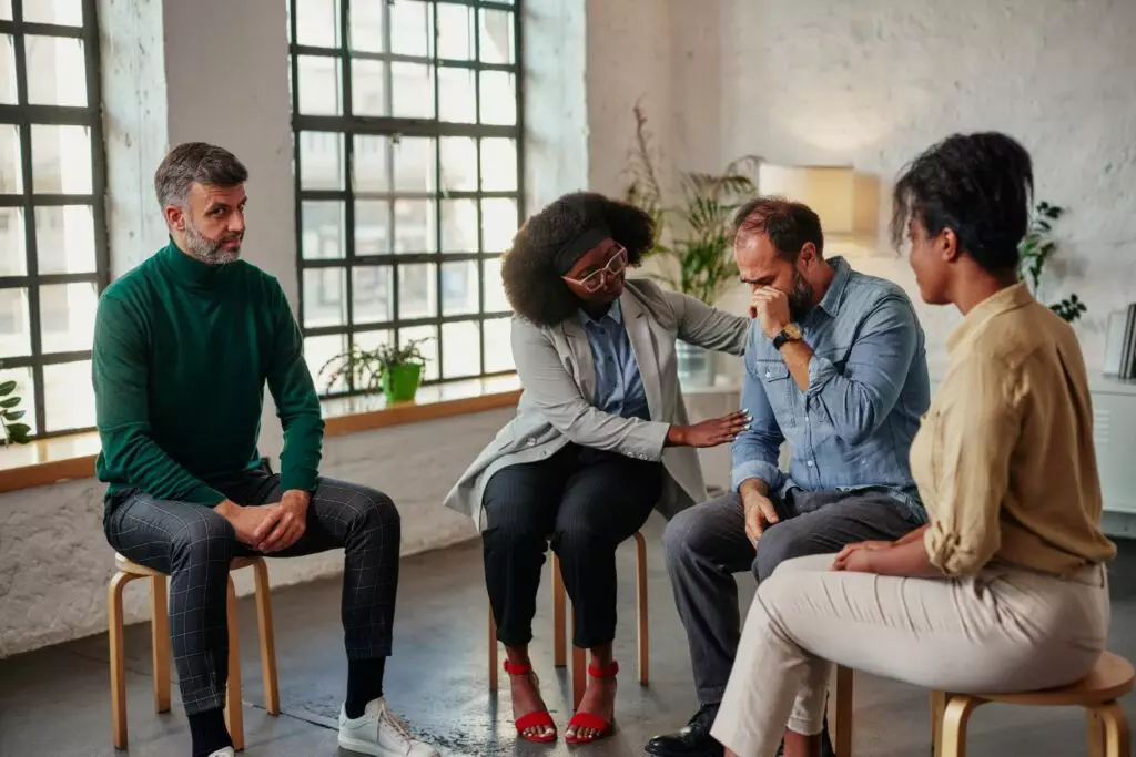 Middle age man sharing struggles during support group meeting with multiracial people siting in circle and comforting him